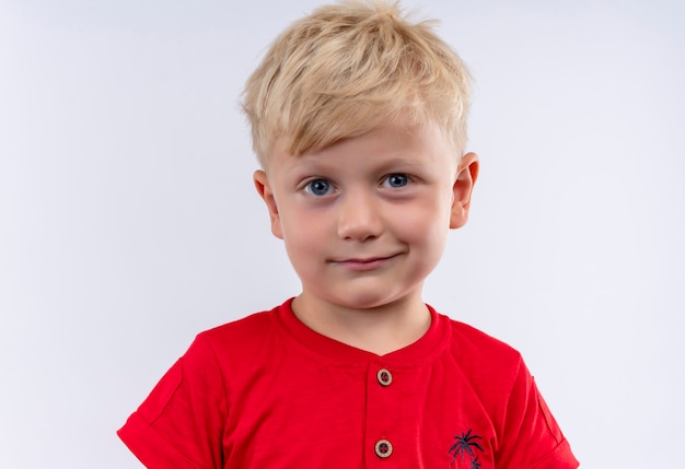 A pretty little boy with blonde hair and blue eyes wearing red t-shirt looking on a white wall