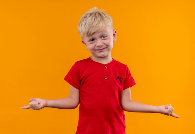 A pretty little boy with blonde hair and blue eyes wearing red t-shirt pointing with index fingers while looking on a yellow wall