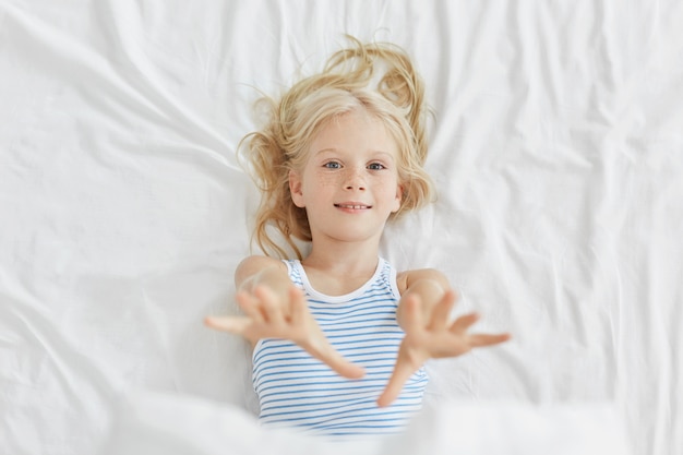 Pretty little child stretching her hands while lying in bed. Blue eyed small girl having rest in bed