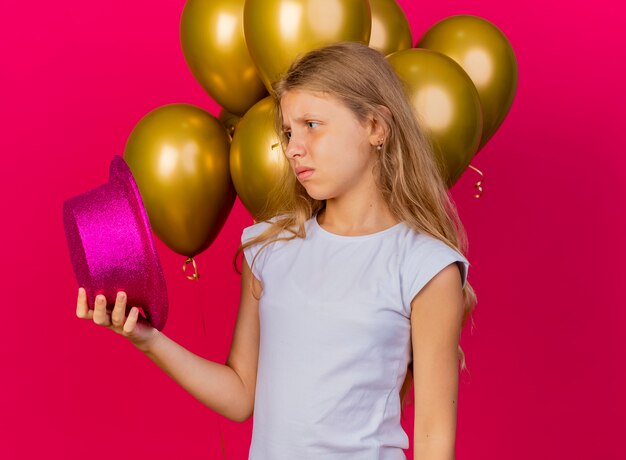 Pretty little girl holding holiday hat with bunch of baloons looking aside with sad expression