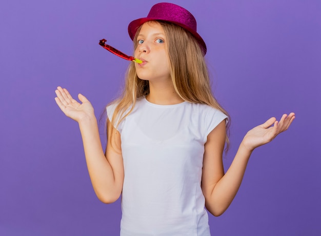 Симпатичная маленькая девочка в праздничной шляпе, дует в свисток, выглядит смущенной, концепция вечеринки по случаю дня рождения стоит на фиолетовом фоне