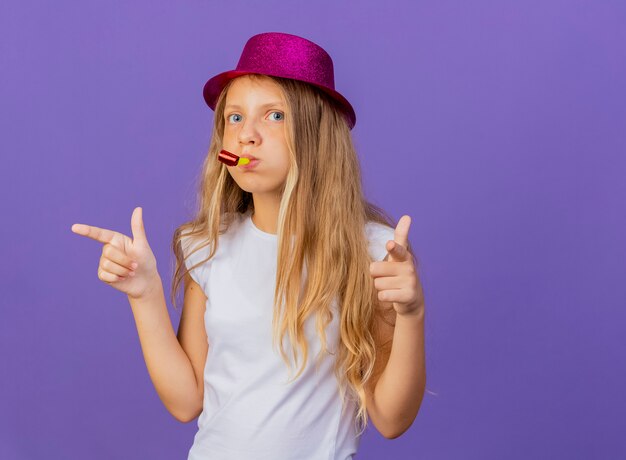Довольно маленькая девочка в праздничной шляпе дует свисток, указывая указательными пальцами на камеру, концепция вечеринки по случаю дня рождения, стоящая на фиолетовом фоне