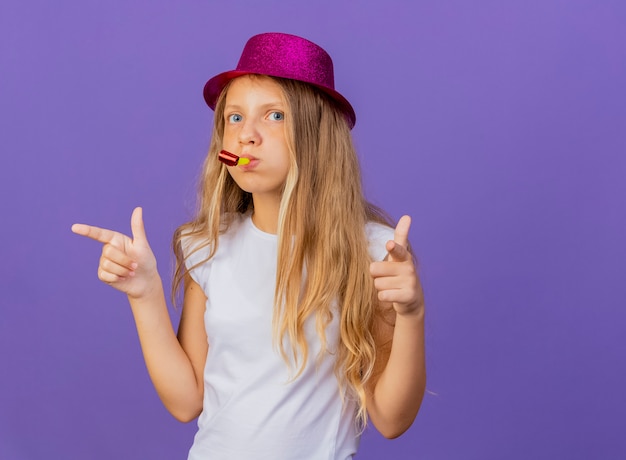 Pretty little girl in holiday hat blowing whistle pointing with index fingers at camera, birthday party concept standing over purple background