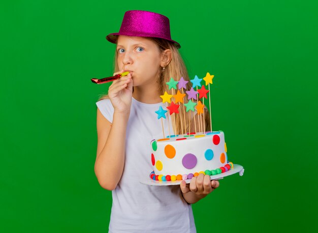 Pretty little girl in holiday hat holding birthday cake blowing whistle