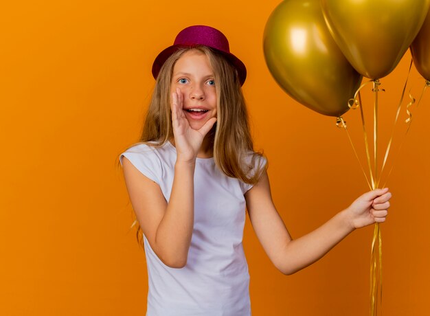 Pretty little girl in holiday hat holding bunch of baloons shouting with hand near mouth, birthday party concept standing over orange background