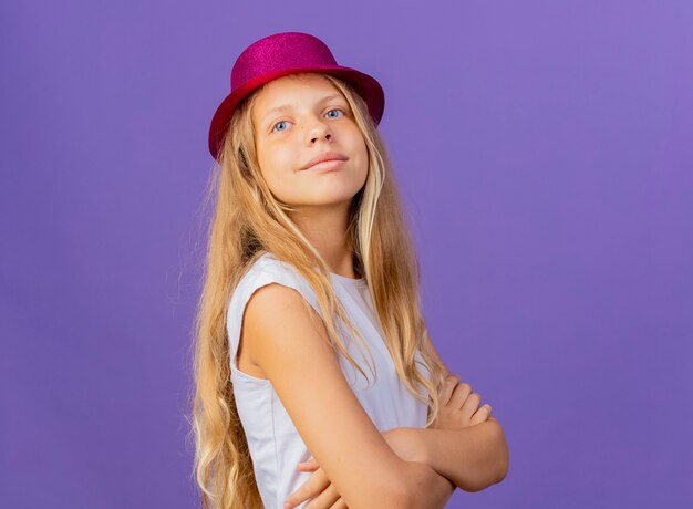 Pretty little girl in holiday hat looking aside with smile on face, birthday party concept standing over purple background