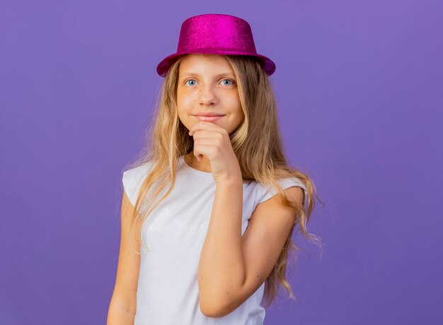 Pretty little girl in holiday hat looking at camera smiling with hand on chin, birthday party concept standing over purple background