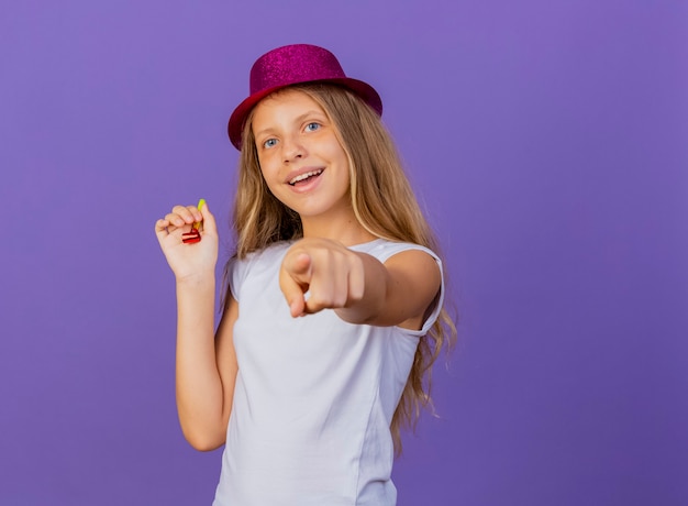Симпатичная маленькая девочка в праздничной шляпе, указывая указательным пальцем на камеру, улыбается счастливым и позитивным, концепция вечеринки по случаю дня рождения стоит на фиолетовом фоне
