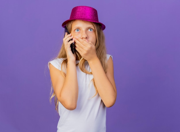 Pretty little girl in holiday hat talking on mobile phone being shocked, birthday party concept standing over purple background