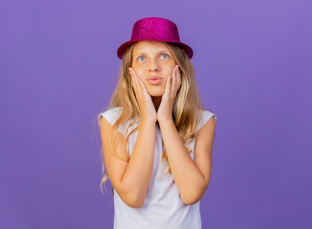 Free photo pretty little girl in holiday hat touching her cheeks feeling positive emotions smiling, birthday party concept standing over purple background