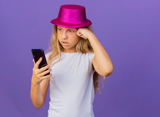 Симпатичная маленькая девочка в праздничной шляпе с помощью смартфона выглядит смущенной и очень обеспокоенной, концепция вечеринки по случаю дня рождения стоит на фиолетовом фоне