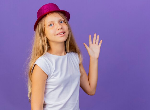 Pretty little girl in holiday hat waving with hand smiling, birthday party concept standing over purple background