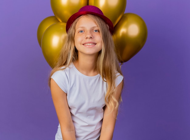 明るい幸せと前向きな笑顔、紫色の背景の上に立っている誕生日パーティーのコンセプトのカメラを見て風船の束と休日の帽子のかわいい女の子