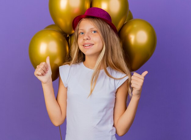 Pretty little girl in holiday hat with bunch of baloons looking at camera smiling showing thumbs up, birthday party concept standing over purple background
