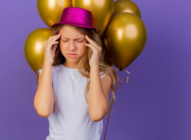 Pretty little girl in holiday hat with bunch of baloons touching her temples having headache, birthday party concept standing over purple background