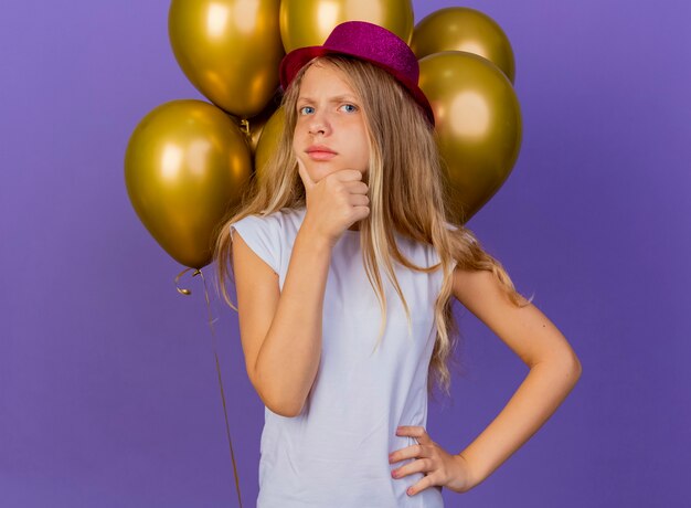 Pretty little girl in holiday hat with bunch of baloons with pensive expression