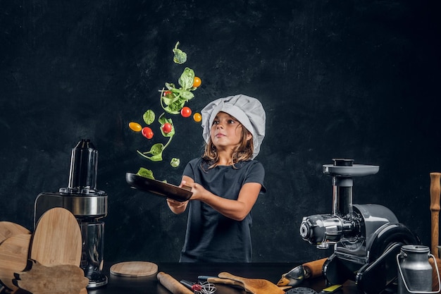 Pretty little girl is tossing vegetables on the pan at dark photo studio.