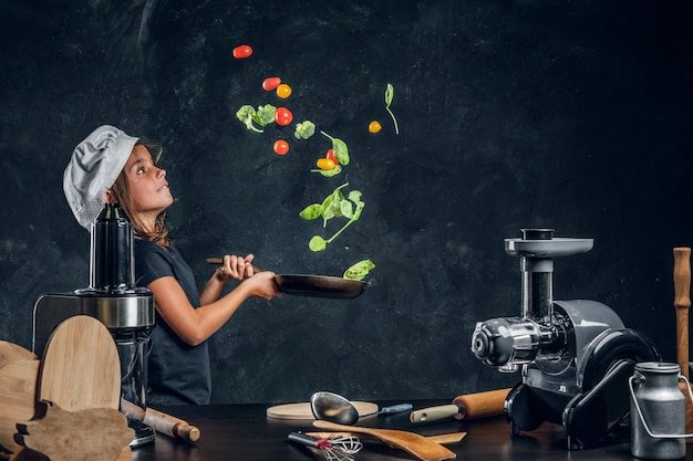 La bambina graziosa sta lanciando le verdure sulla padella allo studio fotografico scuro.