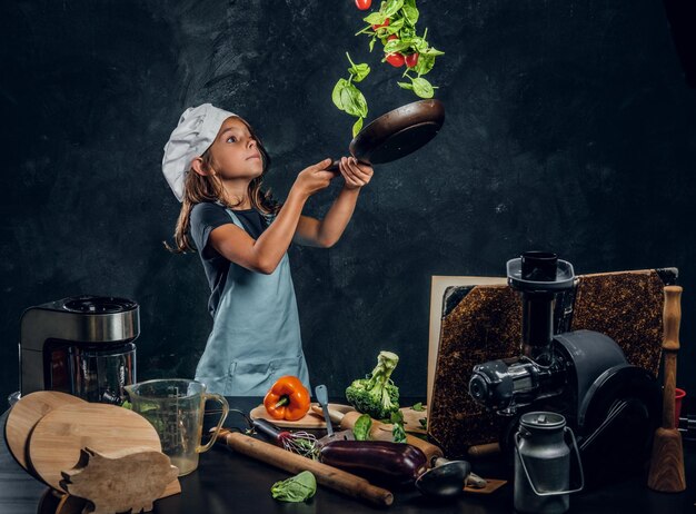 Pretty little girl is tossing vegetables on the pan at dark photo studio.