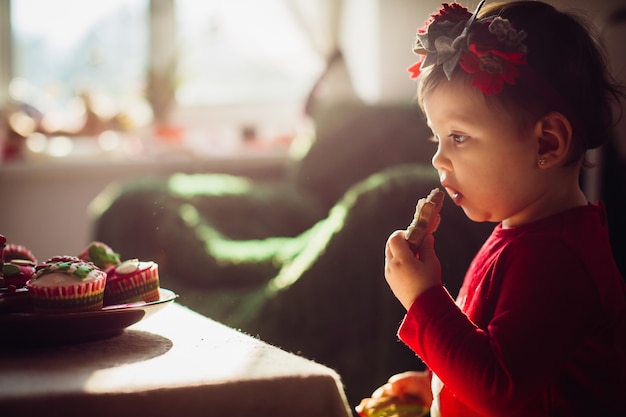 Pretty little girl in red dress eats cookie standing before the table 