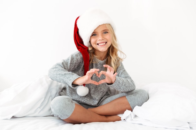 La bambina graziosa in cappello di santa che mostra il cuore firma mentre si siede con le gambe attraversate sul letto bianco