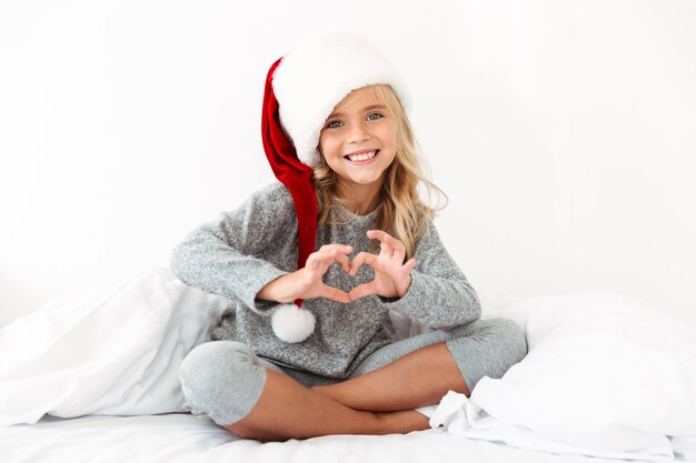 산타의 모자 흰색 침대에 교차 다리와 함께 앉아있는 동안 심장 기호를 보여주는 예쁜 소녀