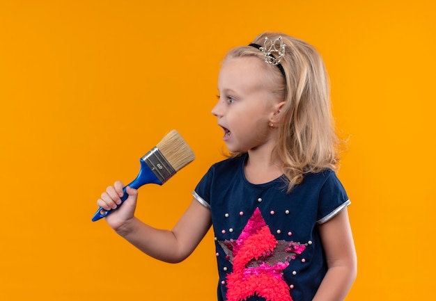 A pretty little girl wearing navy blue shirt in crown headband holding blue paint brush and looking side on an orange wall