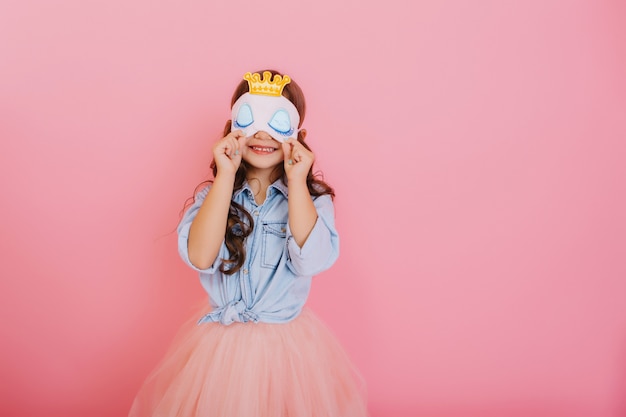Pretty little girl with long brunette hair in tulle skirt holding sleep mask with blue eyes and golden crown isolated on pink background. Celebrating birthday party, having fun at carnival for kids