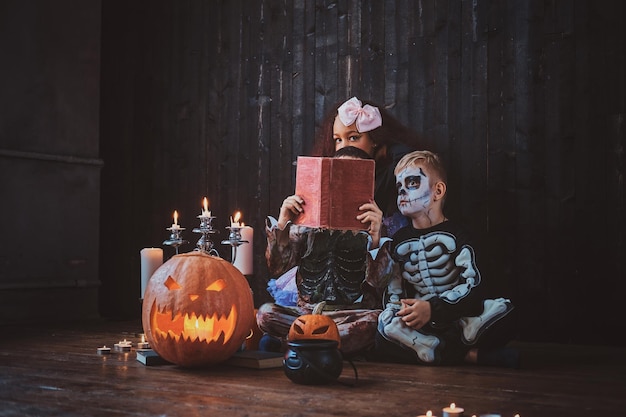 Милые маленькие дети в костюмах на Хэллоуин наслаждаются вечеринкой, читая книгу.