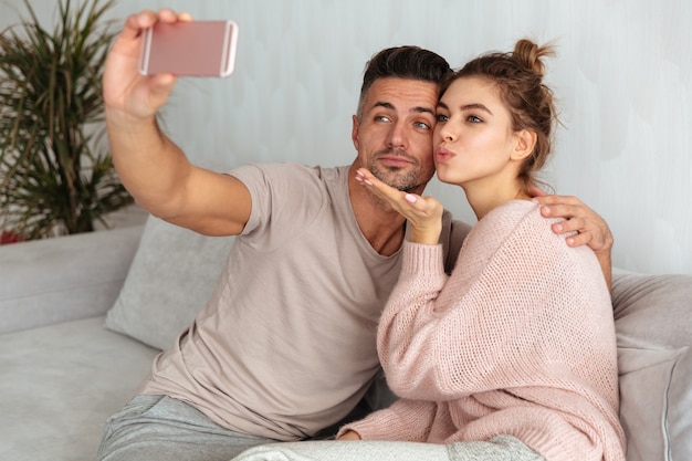 Pretty lovely couple sitting on couch and making selfie on smartphone at home