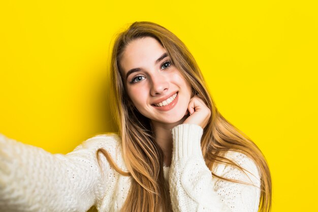 Pretty lovely smiling fashioned girl is making selfie on her phone wearing wide white sweater on yellow