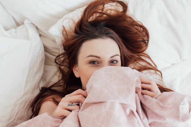 Pretty lovely young woman peeks out of bedsheets covers