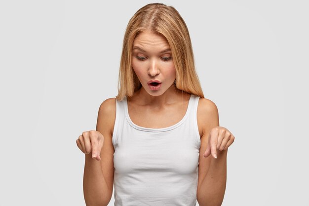 Pretty lovely young woman with long straight hair, points on floor with fore fingers, notices something awful, wears casual white vest