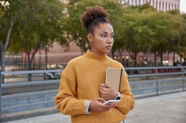 Симпатичная миллениальная девушка с задумчивым выражением лица использует цифровой смартфон и образовательный альбом для рисования, прогуливаясь днем по городу, носит желтый вязаный джемпер, сосредоточенный вдали