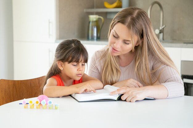 Pretty mom reading book with daughter in kitchen.