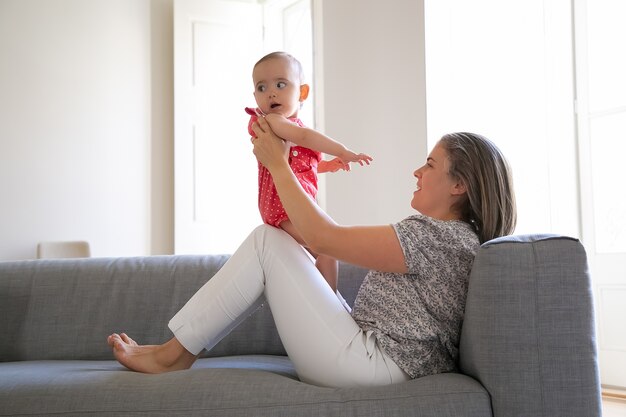 Pretty mother sitting on sofa and holding her little daughter on knees. Adorable baby girl looking away. Long-haired Caucasian mum playing with infant and smiling. Family and motherhood concept