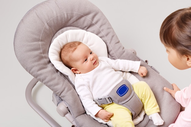 Pretty newborn child in bouncer rocking chair looking with curious expression at her sister