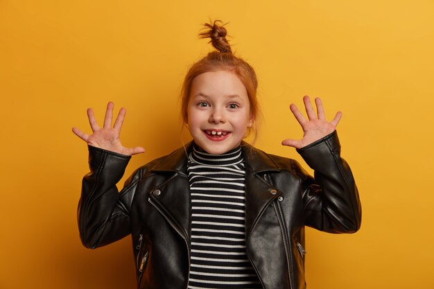 Pretty optimistic girl with foxy hair raises palms, has joyful expression, dressed in striped jumper and leather jacket, being in good mood, waits for parents, isolated over yellow wall