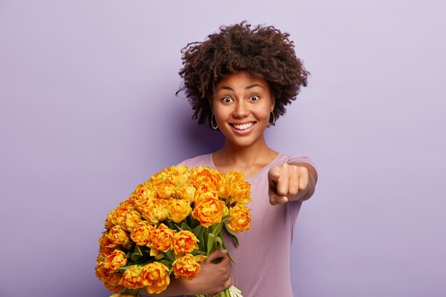 かなり楽観的な女性が前指でまっすぐあなたを指さし、休日に来るように誘い、黄色い花を持って、紫色の壁に隔離されたカジュアルなTシャツを着て、嬉しい笑顔をしています。モノクロ