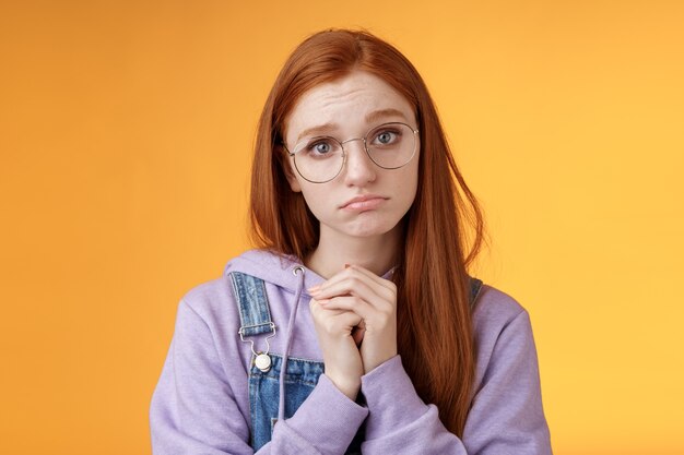 Pretty please. Clingy young sad sulking redhead girl sibling wearing glasses pouting silly press palms begging gesture pleading wanna receive help promise be good ask favour, orange background