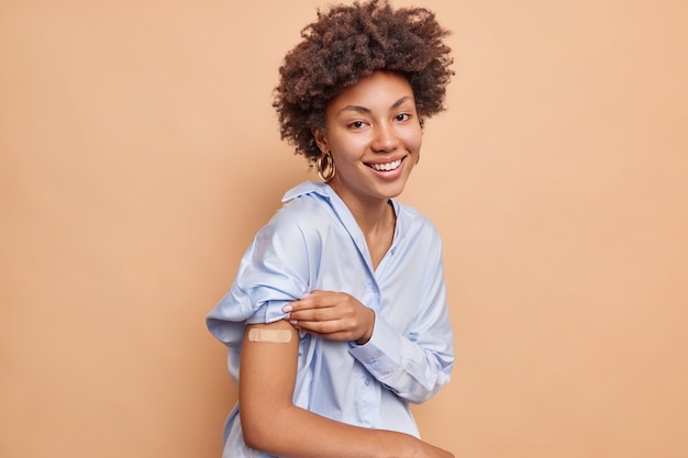 파란색 셔츠를 입은 꽤 기쁘게 생각하는 아프리카계 미국인 여성은 예방 접종을 받은 팔이 베이지색 벽 위에 즐겁게 격리된 접착 석고 미소를 보여줍니다.