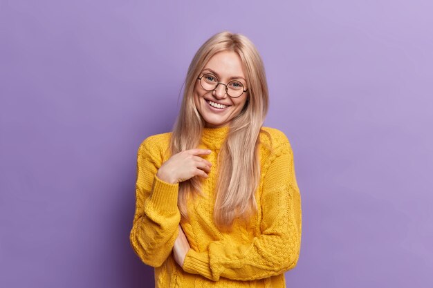 Pretty pleased European woman with blue eyes and blonde hair smiles pleasantly shows white teeth has pleasant talk with someone happy to hear good news wears big optical glasses poses indoor