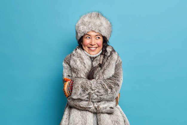 Довольно довольная молодая эскимосская женщина, обнимая себя, чувствует себя комфортно в зимнем пальто, а шляпа с мечтательным выражением лица изолирована от синей стены