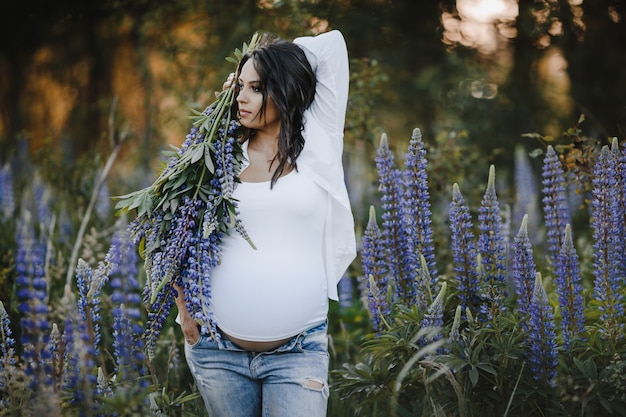 Pretty pregnant woman stands with lupines bouquet among field