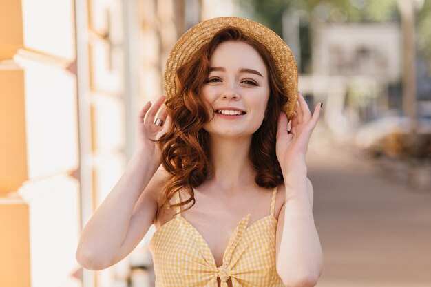 Pretty red-haired girl with cute face expression smiling on city. Outdoor shot of carefree curly female model enjoying summer walk.