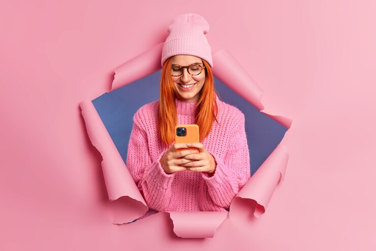 無料Photo かなり赤毛の女性がスマートフォンのデバイスを喜んで見て、スタイリッシュな服を着た彼女のメールの笑顔をチェックします。