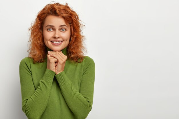 Pretty redhead woman looks with lovely expression, has seductive look, presses hands together near chin, happy to live carefree life, wears casual green jumper, stands against white wall, empty space