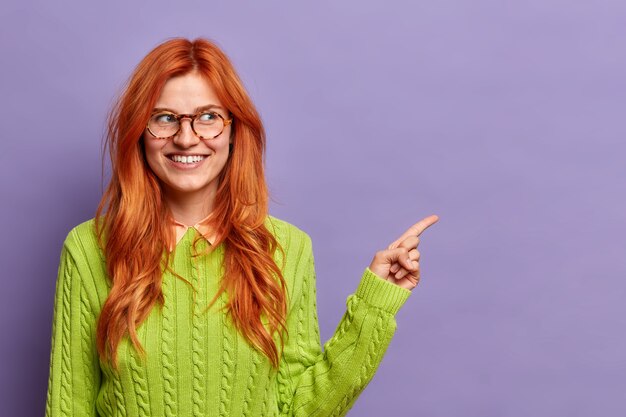 Pretty redhead woman with toothy smile points away on copy space