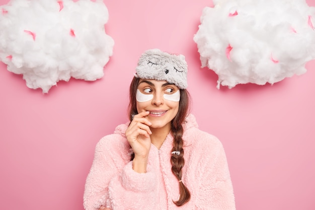 Pretty satisfied European girl smiles gently has curious face expression enjoys comfort and serene atmosphere at home wears sleepmask with pajama isolated over pink wall going to sleep