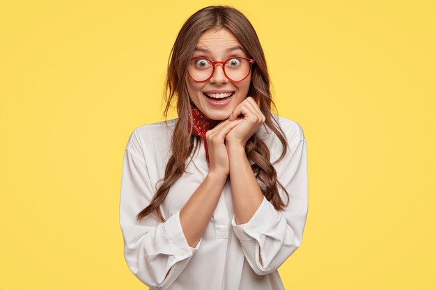 Pretty satisfied lady expresses good feelings, keeps hands together under chin, has happy look, wears oversized white shirt, expresses like, isolated over yellow wall. People and joy concept